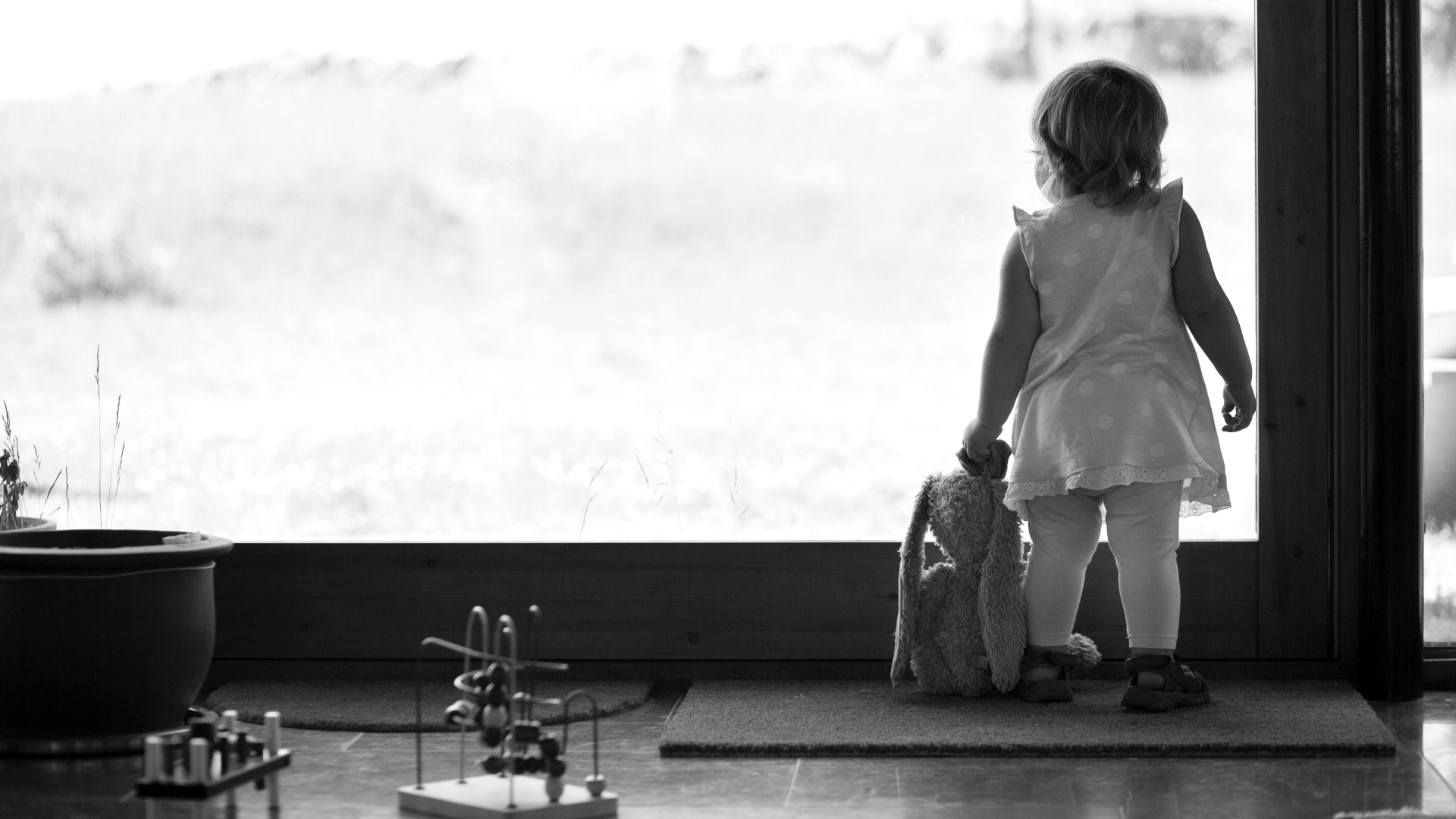 Egy 18-24 hónap közötti kislány néz ki az ablakon, kezében plüssnyúl