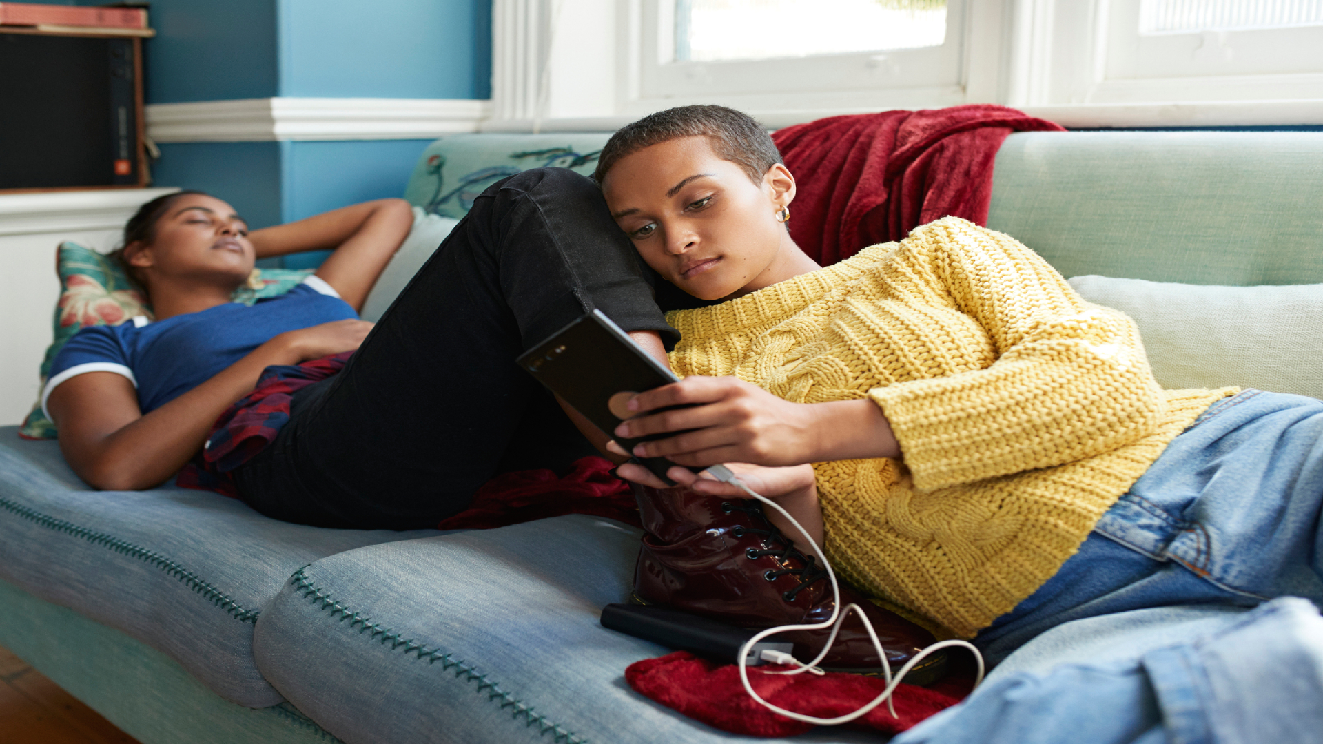 Kanapén fekvő fiatalok, egyikük alszik, a másik telefonozik.