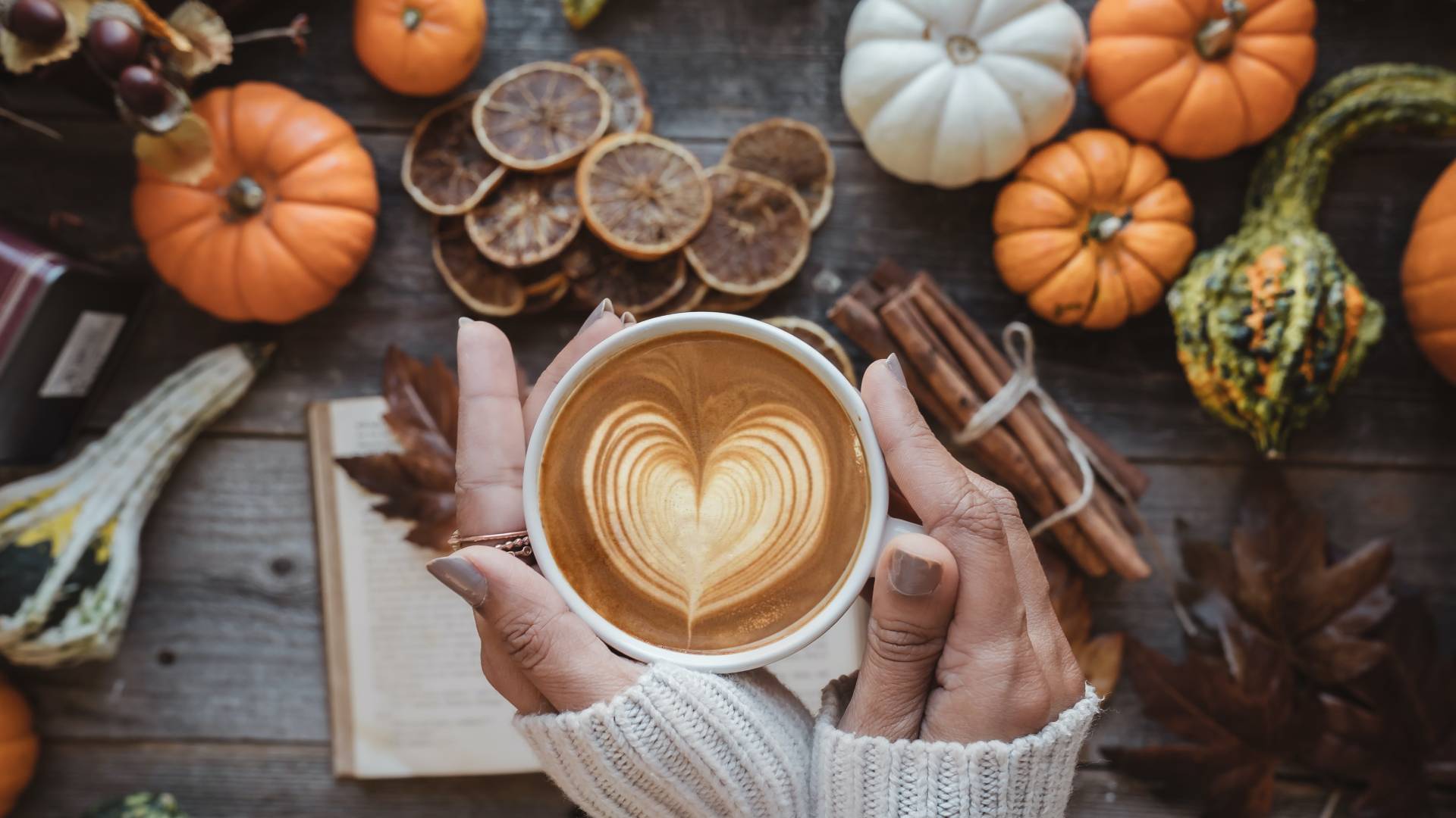 Kávét tartó női kéz, háttérben könyv és tökök.