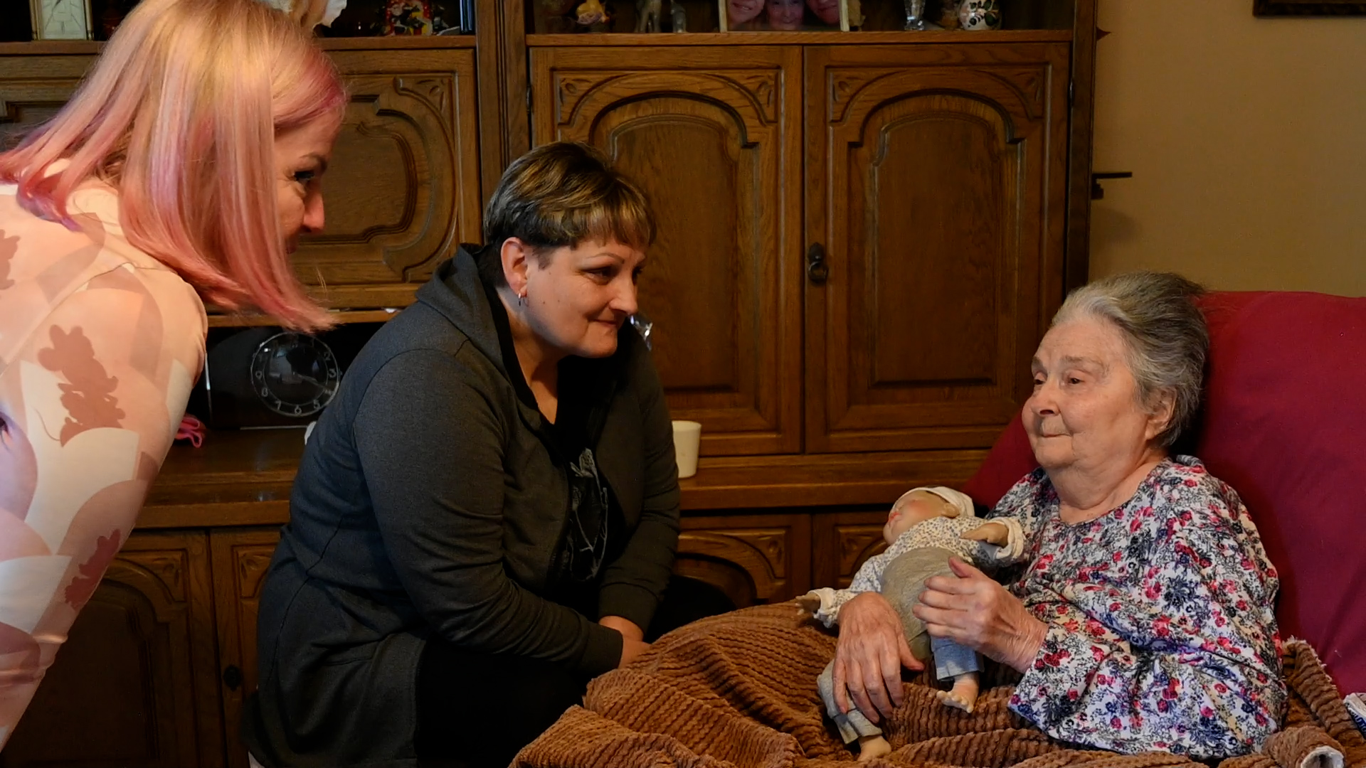 idős hölyg babával ül, két nő nézi