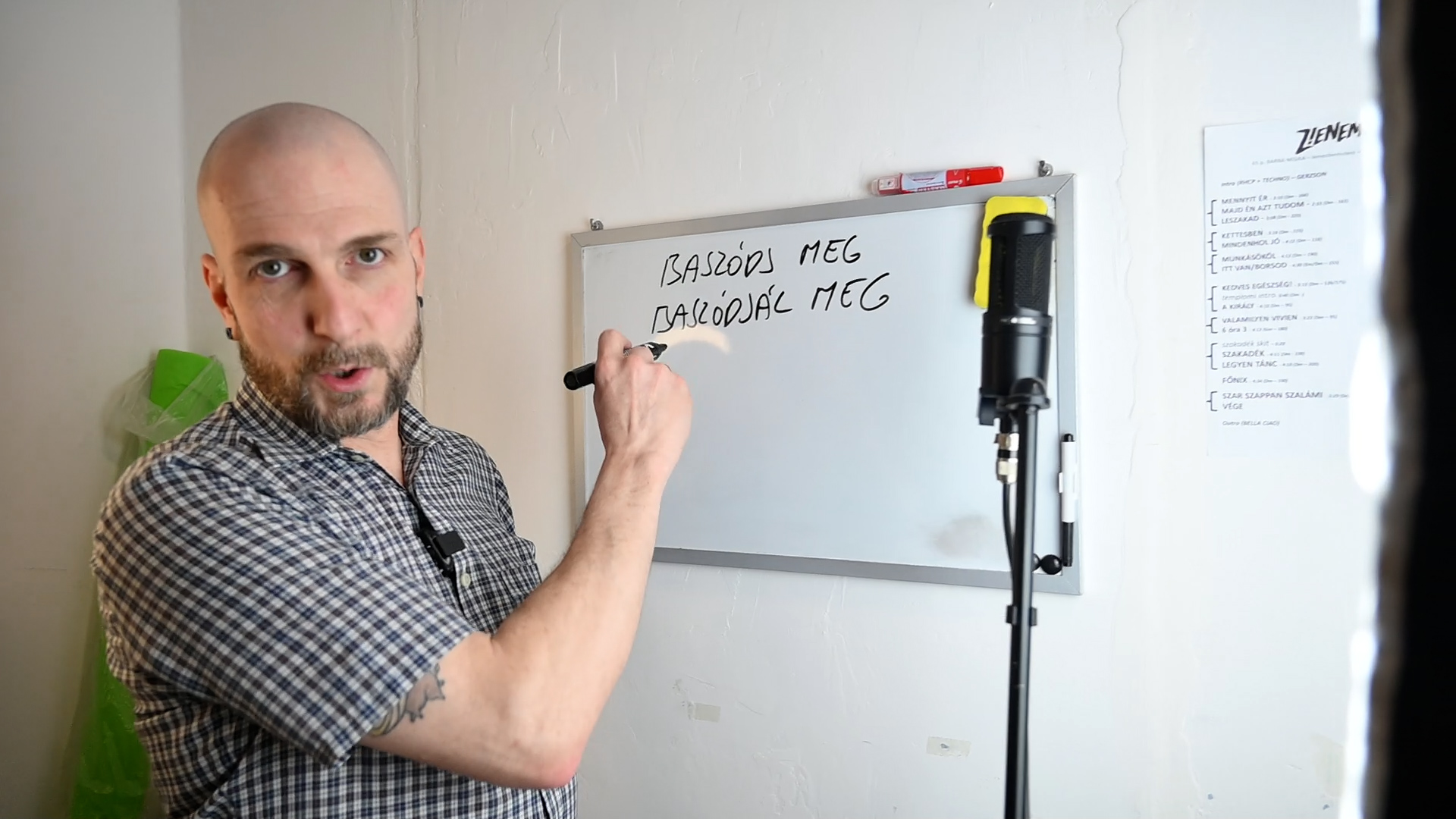 kopasz férfi egy táblára ír, és a kamerába néz